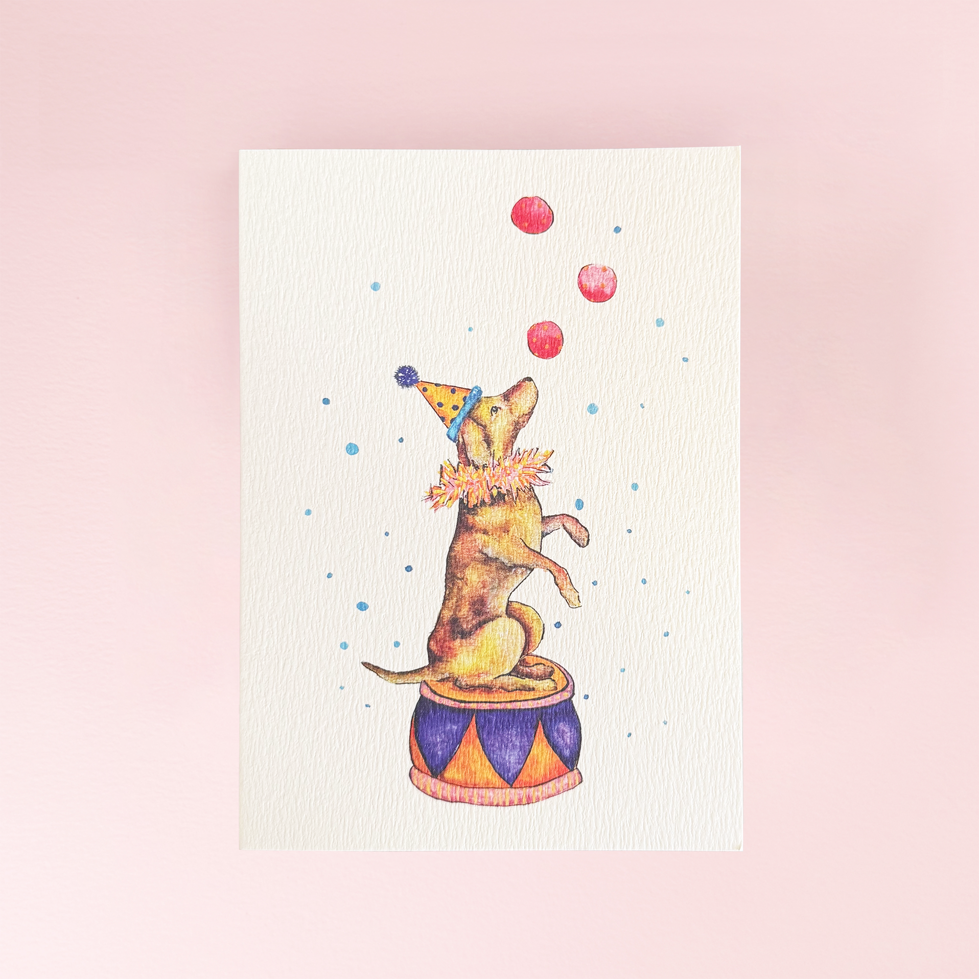 A vertical greeting card featuring a watercolour illustration of a small dog balancing on a circus drum while juggling red balls, set against a cream background
