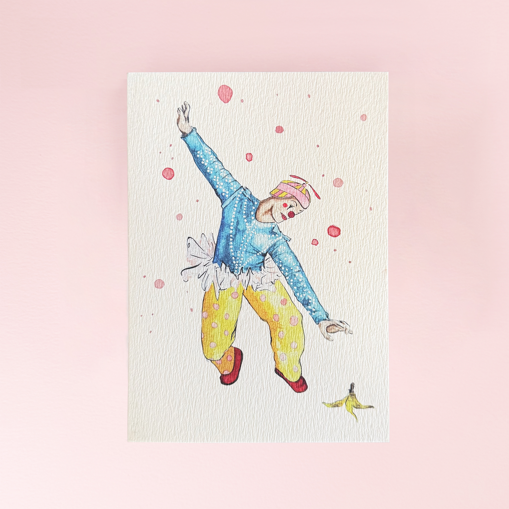 A greeting card showing a performing circus entertainer in a colourful outfit and a banana peel on the floor. Ona cream card background