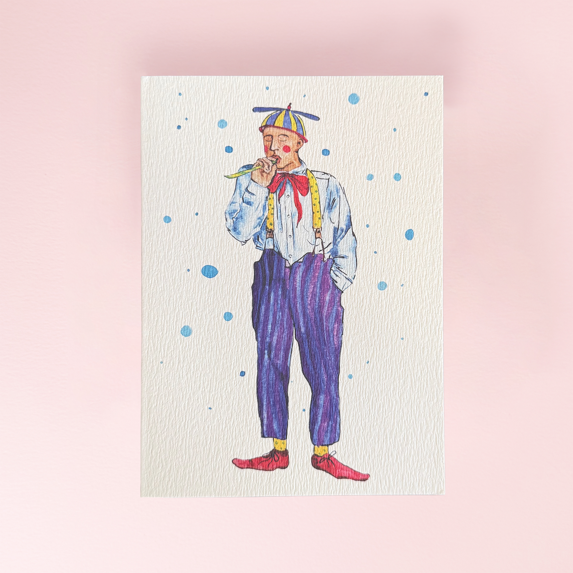 A greeting card showing a watercolour painting of a circus entertainer in a colourful outfit with blue polka dots scattered around him on a cream background
