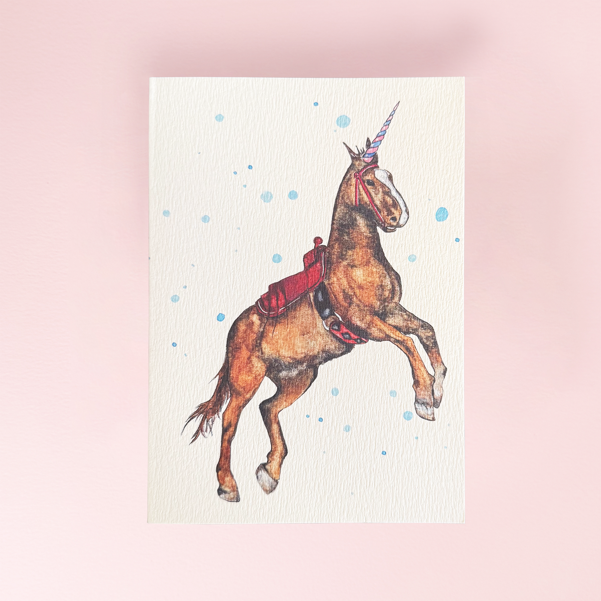 A vertical art greeting card featuring a brown unicorn with a red saddle and horn, set against a light cream background with blue watercolor splatters, displayed on a soft pink background.