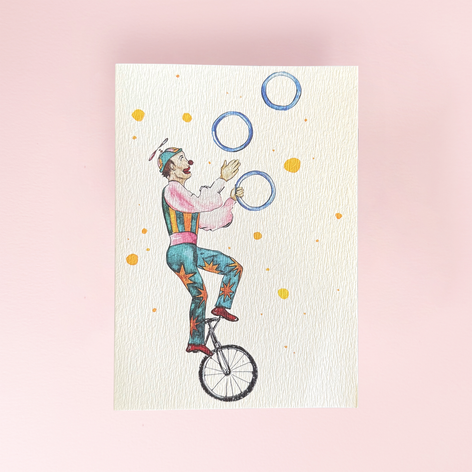 A greeting card showing a juggler on a unicycle in a colourful outfit on a cream card background.